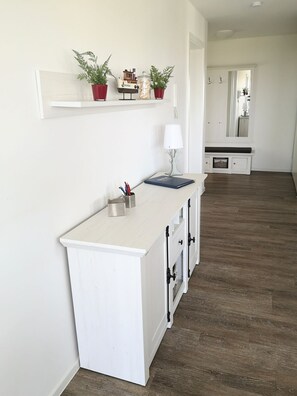 Cocina blanca e impecable con encimera de madera, un lavamanos pequeño y un armario con cajones. Encima de la encimera hay una estantería con objetos decorativos y plantas.