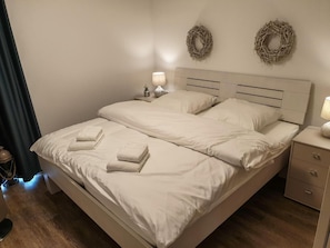 A neatly made bed with two pillows and two folded towels, a bedside table with a lamp, and two wall-mounted wreaths. - 47.5.S Waterkant - Cozy, high-quality vacation apartment, own sauna, south-facing, (Wangerland)