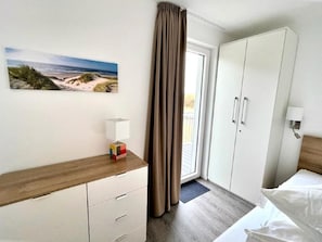 Ett hotellrum med en säng, en garderob, ett nattduksbord med en lampa, en tavla med ett strandmotiv och ett fönster med gardiner.