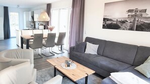 A modern living room with a grey sofa, white armchairs, and a wooden coffee table. A dining area with a table and chairs is visible in the background. - House No. 22, Ferienhaus Deichgraf - Modern vacation home, fireplace, sauna, south (Wangerland)