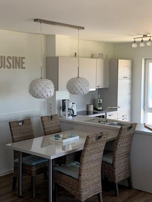 Une cuisine moderne avec des meubles blancs, une table à manger entourée de chaises en osier et des luminaires suspendus.
