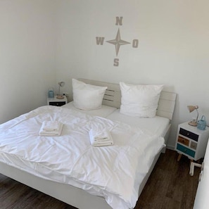 Un lit bien fait, avec du linge de lit blanc et deux serviettes pliées, une table de chevet blanche avec une lampe, et une boussole murale.