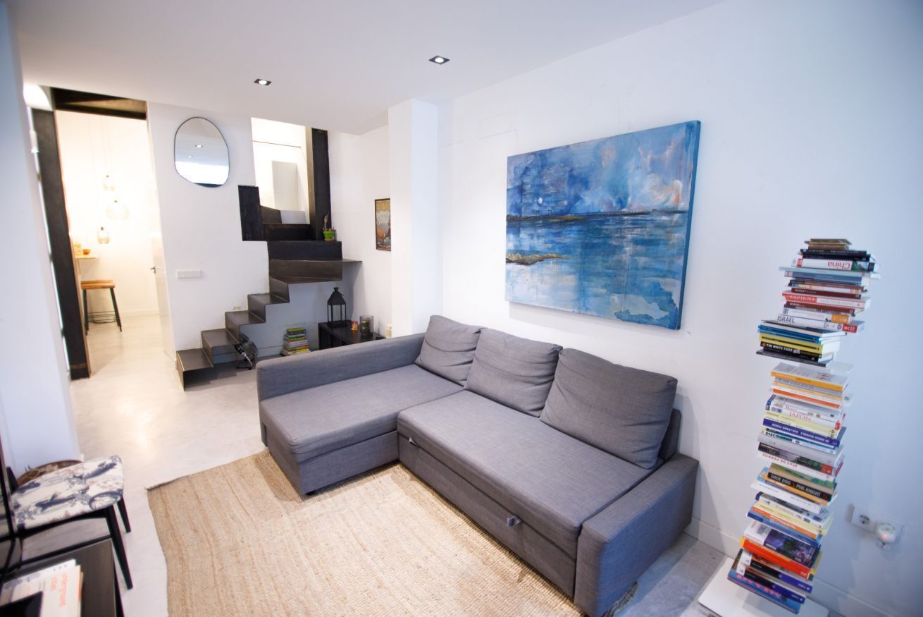 A modern living room with a grey sectional sofa, a staircase, a large abstract painting, and a bookshelf filled with books.