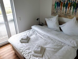 A hotel room with a bed, white linens, two towels, a painting on the wall, and a view through a sliding door. - 47.7 Mehrblick - Luxury vacation apartment, own sauna, large terrace, sea view (Wangerland)