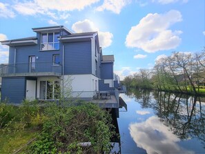 En modern tvåvåningsbyggnad med en balkong, belägen intill ett lugnt vattendrag som speglar himlen och träden.