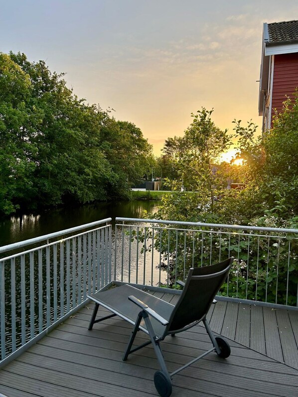 A deck with a chair overlooking a river, surrounded by trees and a building.