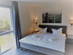 Une chambre d’hôtel moderne avec un grand lit, une table de chevet, une lampe fixée au mur et une fenêtre donnant sur un paysage verdoyant.