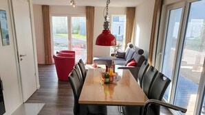 A modern dining area with a wooden table, black chairs, and a red pendant light. A red chair is visible in the background, and a large window offers a view of the outside. - House no. 07, Ferienhaus Deichgraf - Modern vacation home, sauna, south-facing terrace, (Wangerland)
