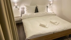 A neatly made bed with white linens and two folded towels, a bedside table with a lamp, and a wall-mounted light fixture. - House no. 07, Ferienhaus Deichgraf - Modern vacation home, sauna, south-facing terrace, (Wangerland)