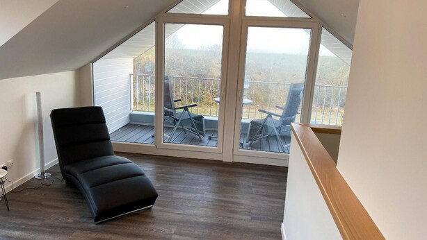 A spacious room with a sloped ceiling, a black leather chair, and a large sliding glass door leading to an outdoor deck with chairs. - House no. 07, Ferienhaus Deichgraf - Modern vacation home, sauna, south-facing terrace, (Wangerland)