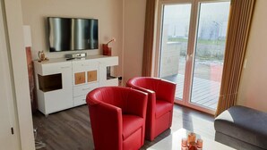 A modern living room with a flat-screen TV, a white entertainment unit, two red chairs, a grey sofa, and a large sliding glass door leading to an outdoor deck. - House no. 07, Ferienhaus Deichgraf - Modern vacation home, sauna, south-facing terrace, (Wangerland)