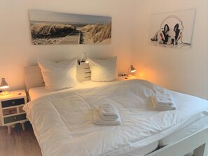 Ein ordentlich bezogenes Bett mit weißen Leinentüchern und zwei gefalteten Handtüchern, eine Nachttischlampe, ein Nachttisch und ein an der Wand hängendes Bild mit einer Strandansicht.