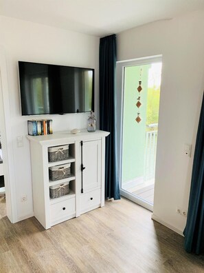 A room with a flat-screen TV mounted above a white cabinet with shelves, a sliding glass door leading to an outdoor area, and a wooden floor. - 47.1.S Waterkant - Cozy, well-kept vacation apartment near the beach, own (Wangerland)