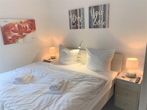A neatly made bed with white linens and towels, two bedside lamps, a nightstand with a remote control, and two wall art pieces with text. - 47.1.S Waterkant - Cozy, well-kept vacation apartment near the beach, own (Wangerland)