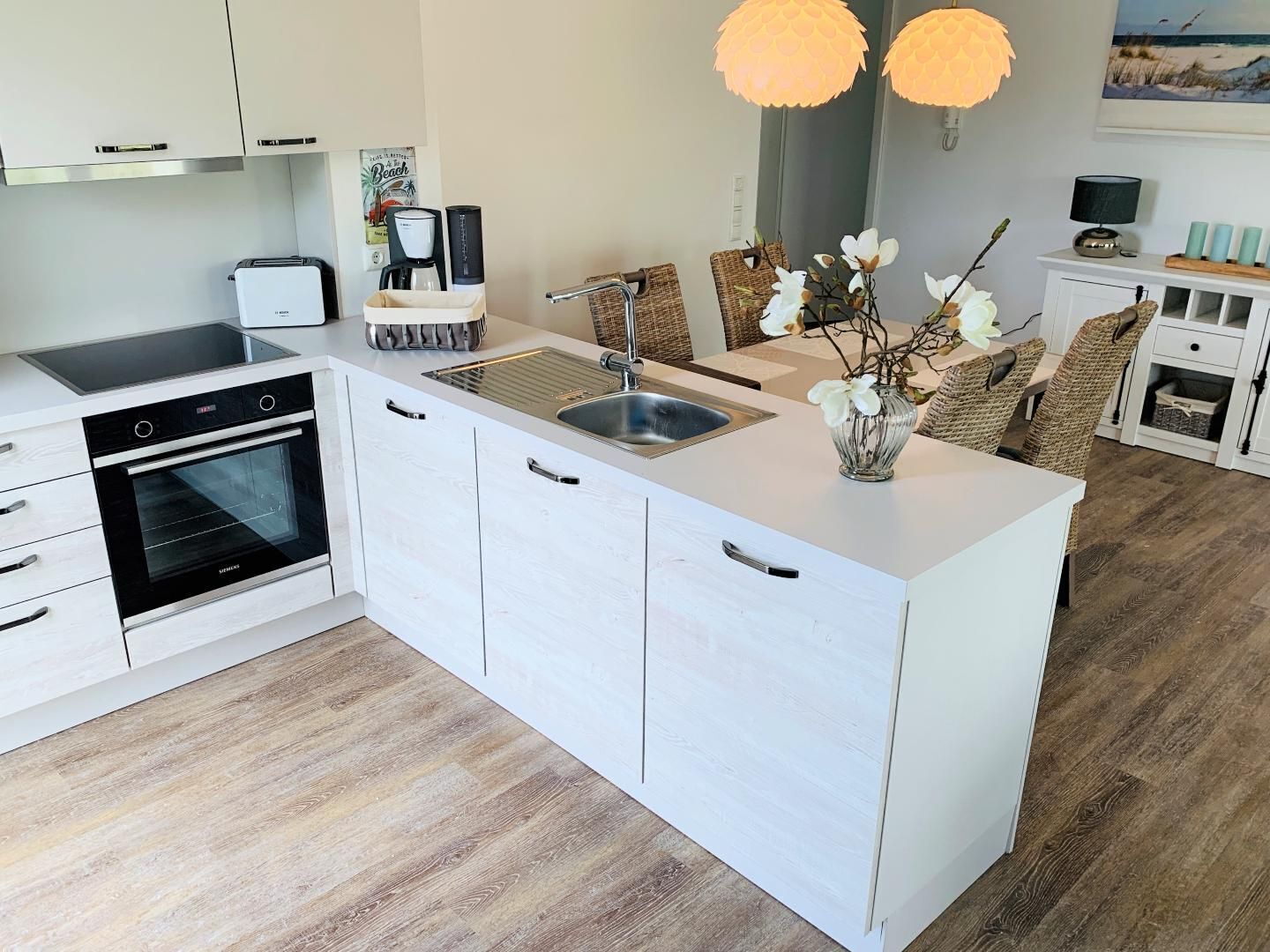 A modern kitchen with white cabinetry, a built-in oven, a sink, and a coffee maker. There are wicker chairs and a vase with flowers on the island.