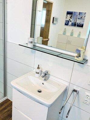 Ein Badezimmer mit einem weißen Waschbecken, einer Glasablage darüber und einem Spiegel, der den Raum widerspiegelt.