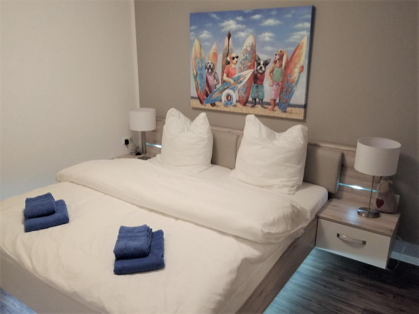Quarto de hotel com uma cama, duas luminárias de cabeceira, um quadro na parede e piso de madeira.