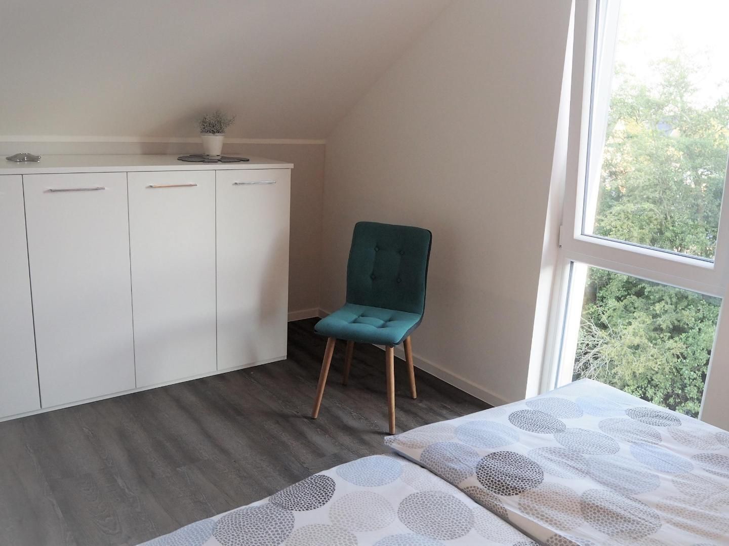 Una camera da letto con un letto, una poltrona color petrolio, un armadio e una finestra vista alberi.