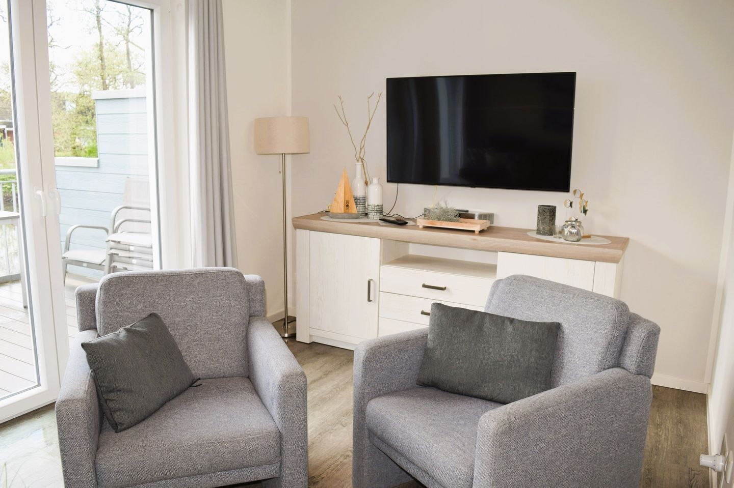 Un salon moderne avec deux fauteuils gris, un téléviseur à écran plat fixé sur une console en bois clair et un lampadaire.