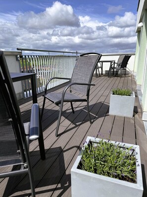 Eine Terrasse mit Stühlen und Tisch, umgeben von einer Brüstung, mit Blick auf ein Gewässer.
