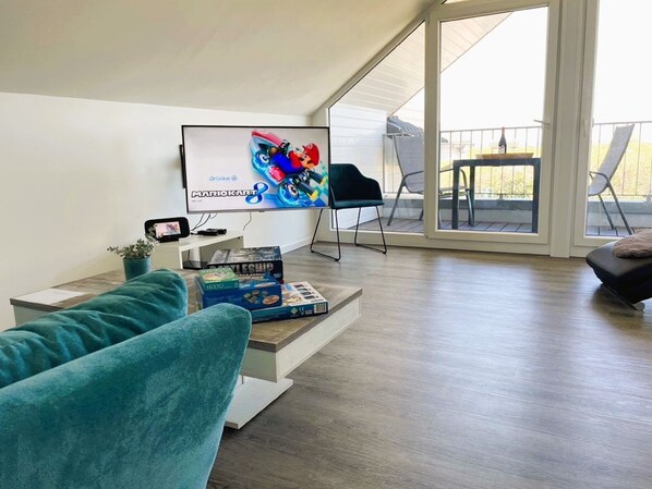 Ein modernes Wohnzimmer mit einem großen Flachbildfernseher, einem türkisfarbenen Sofa und einem Couchtisch mit Gesellschaftsspielen.