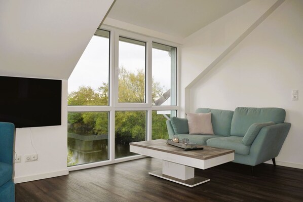 Ein modernes Wohnzimmer mit einem großen Fenster, einem Flachbildfernseher, einem türkisfarbenen Sofa und einem Couchtisch.