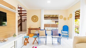 A modern living room with a TV, blue and white chairs, a sofa, and a staircase. - GB45 Excelente Casa 4 Suítes - Guarajuba (Camaçari)