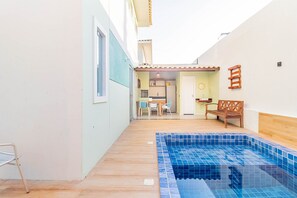 A swimming pool with tiled edges, a wooden deck, a dining area with chairs and a table, and a kitchen visible through an open door. - GB45 Excelente Casa 4 Suítes - Guarajuba (Camaçari)