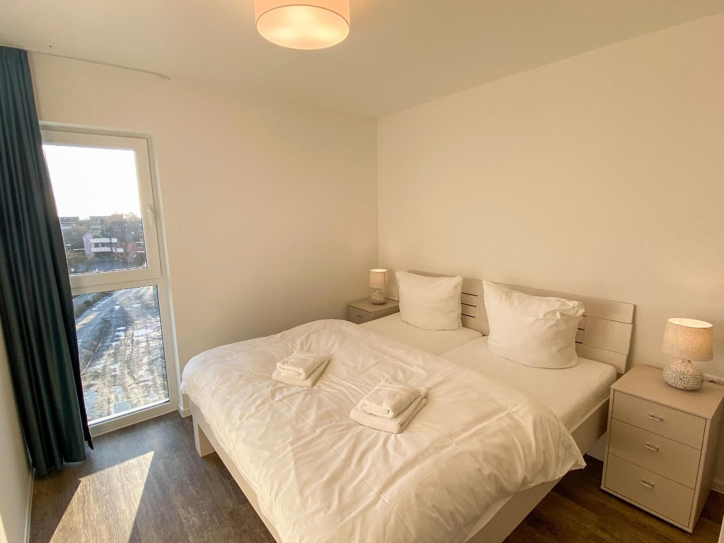 A bedroom with a large bed, two bedside tables, a window with curtains, and a view of the city.