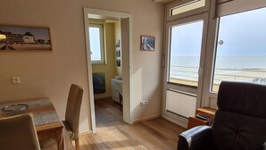 A room with a wooden floor, a dining table with chairs, a large window with a view of the sea, and a door leading to another room.