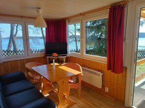 A cozy cabin room with a wooden interior, a dining table with chairs, a sofa, a TV, and large windows with curtains.