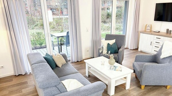 A modern living room with a grey sofa, a white coffee table, and a grey armchair. There is a dining table and chairs outside the window, and a flat-screen TV mounted on the wall.