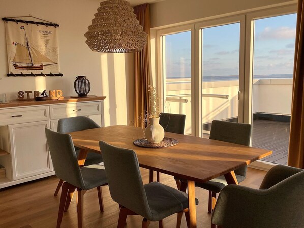 Uma sala de jantar com uma mesa e cadeiras de madeira, um lustre pendente, um vaso decorativo e vista para o mar através de grandes janelas.