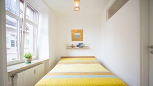 A narrow room with a bed, a window, a radiator, and a shelf with books and a plant.