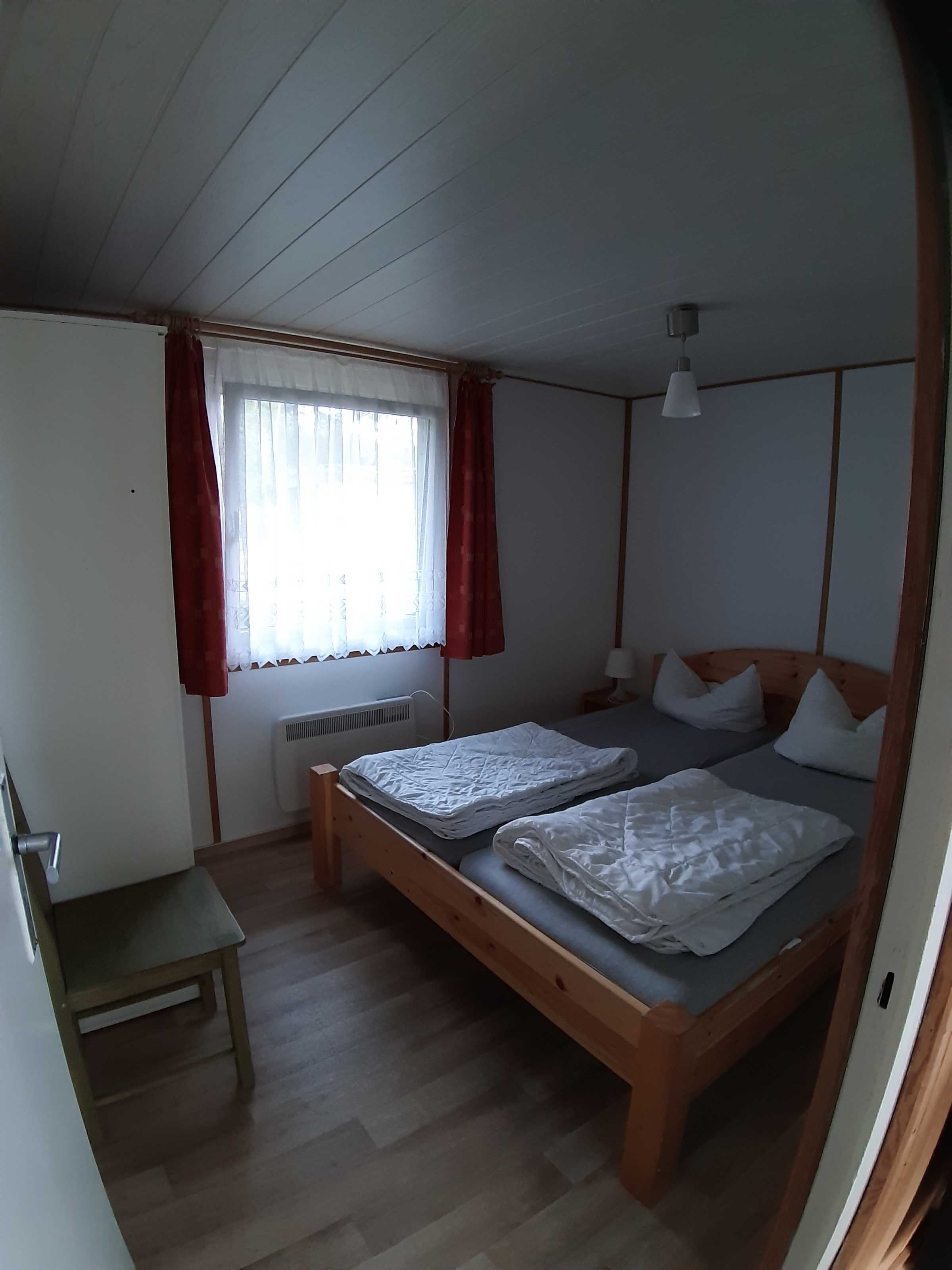 A small, single-room accommodation with two wooden beds, a small table, and a window with curtains.