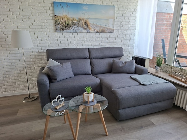Un canapé gris avec des coussins, une table basse en verre ornée de deux plantes en pot et un tableau représentant une scène de plage sur un mur de briques blanches.