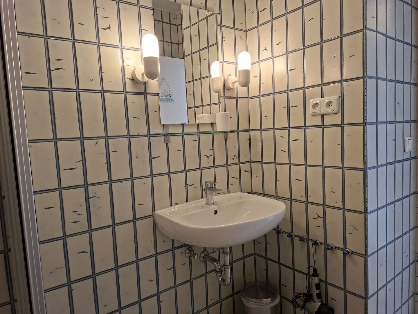 A bathroom with a grid-patterned tile wall, a white sink, and two wall-mounted light fixtures.