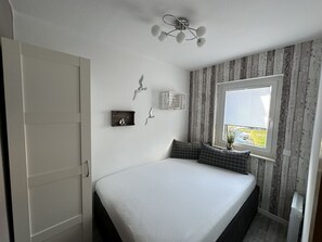 A bedroom with a bed, a window with blinds, a ceiling fan, and a wall-mounted shelf with decorative items.