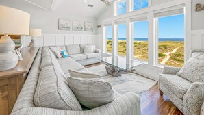Ein geräumiges Wohnzimmer mit großem Fenster zum Strand, Holzboden und einem modernen Deckenventilator.