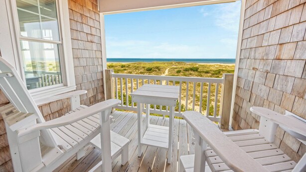 Eine Holzterrasse mit weißen Möbeln, Blick auf den Strand und einem klaren Himmel.