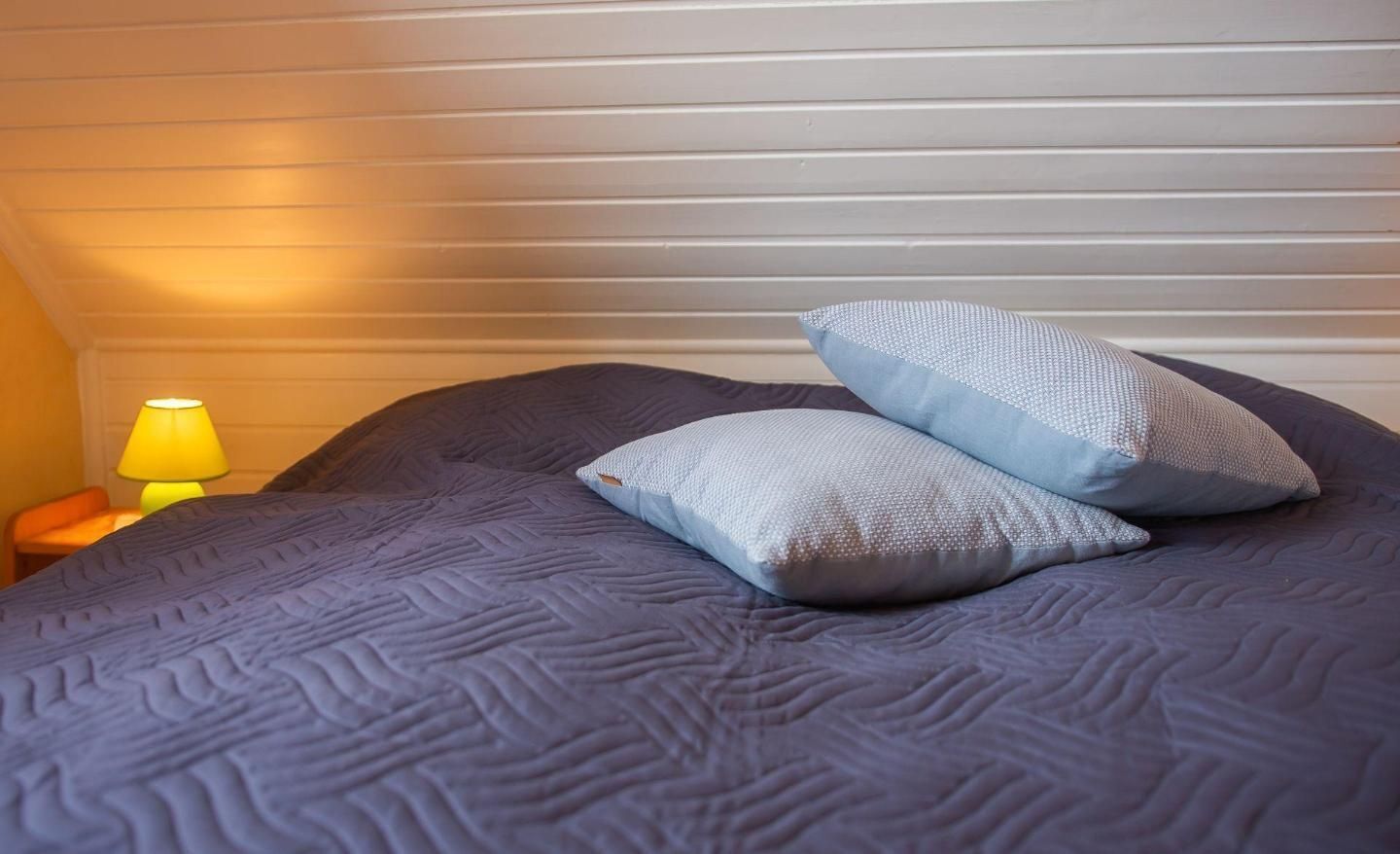 Un lit recouvert d’une couverture bleue à motifs, avec deux oreillers, une lampe jaune posée sur une table de chevet, et une fenêtre avec des stores horizontaux.