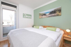 A bedroom with a large bed, a view of the outdoors through a sliding door, a wall-mounted artwork, and a bedside table with a lamp. - Lindenhof - Fresh breeze - Charming apartment, close to the beach! (Dahme)