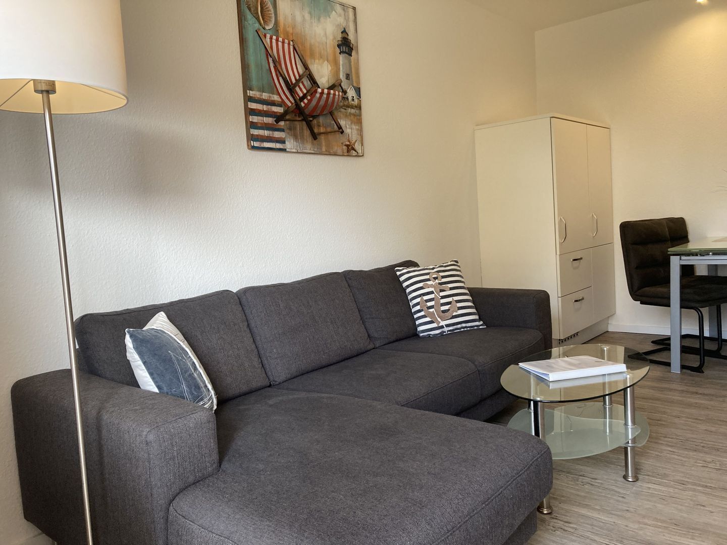 Uma sala de estar moderna com um sofá seccional cinza, uma mesa de centro de vidro, um abajur, um quadro de um farol e um armário branco.