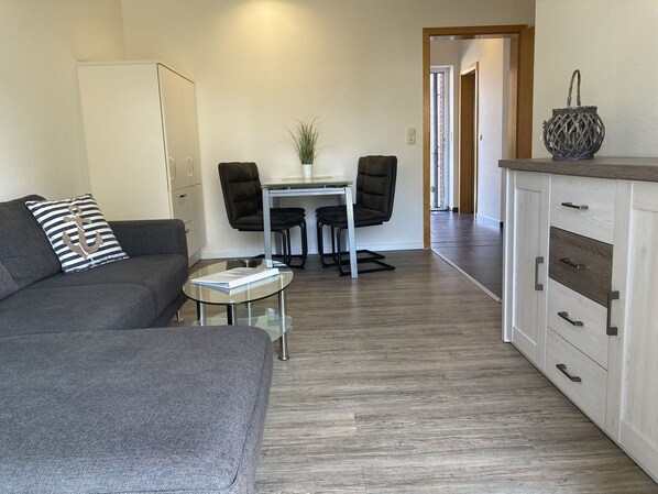 A modern living room with a grey sofa, a glass coffee table, a dining table with two chairs, and a kitchen area with cabinets and a countertop. - The "Seepferdchen" in Haus Meeresglück, close to the beach and central! - House (Dahme)