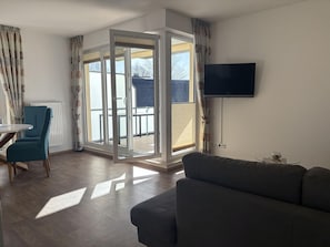 Un salon moderne avec un canapé gris, un fauteuil turquoise, une petite table ronde et un téléviseur fixé au mur.