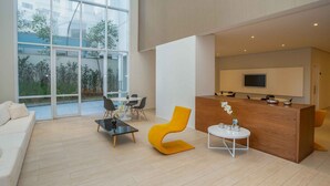 A modern living room with a sofa, a coffee table, a yellow chair, and a reception desk with a TV. - Urbe Bela Vista (São Paulo)