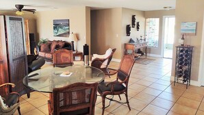 A spacious living area with a glass-top dining table surrounded by wicker chairs, a sofa, and a coffee table. There is a ceiling fan, a fireplace, and a wine rack with bottles.