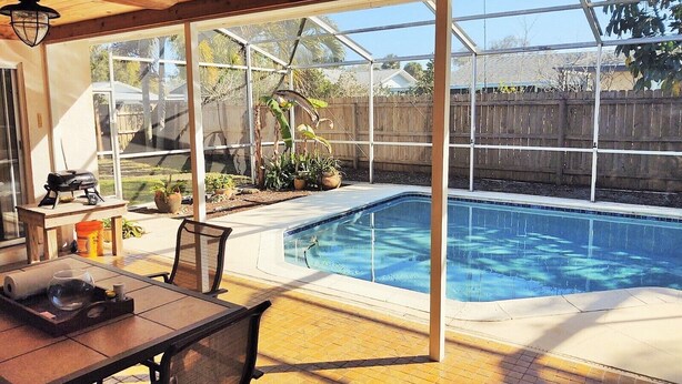 A backyard with a swimming pool, a wooden deck, a table with outdoor seating, and a grill.
