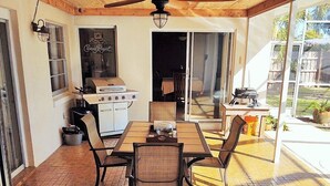 A patio with a brick floor, a wooden ceiling, and a ceiling fan. There is a table with chairs and a grill outside.
