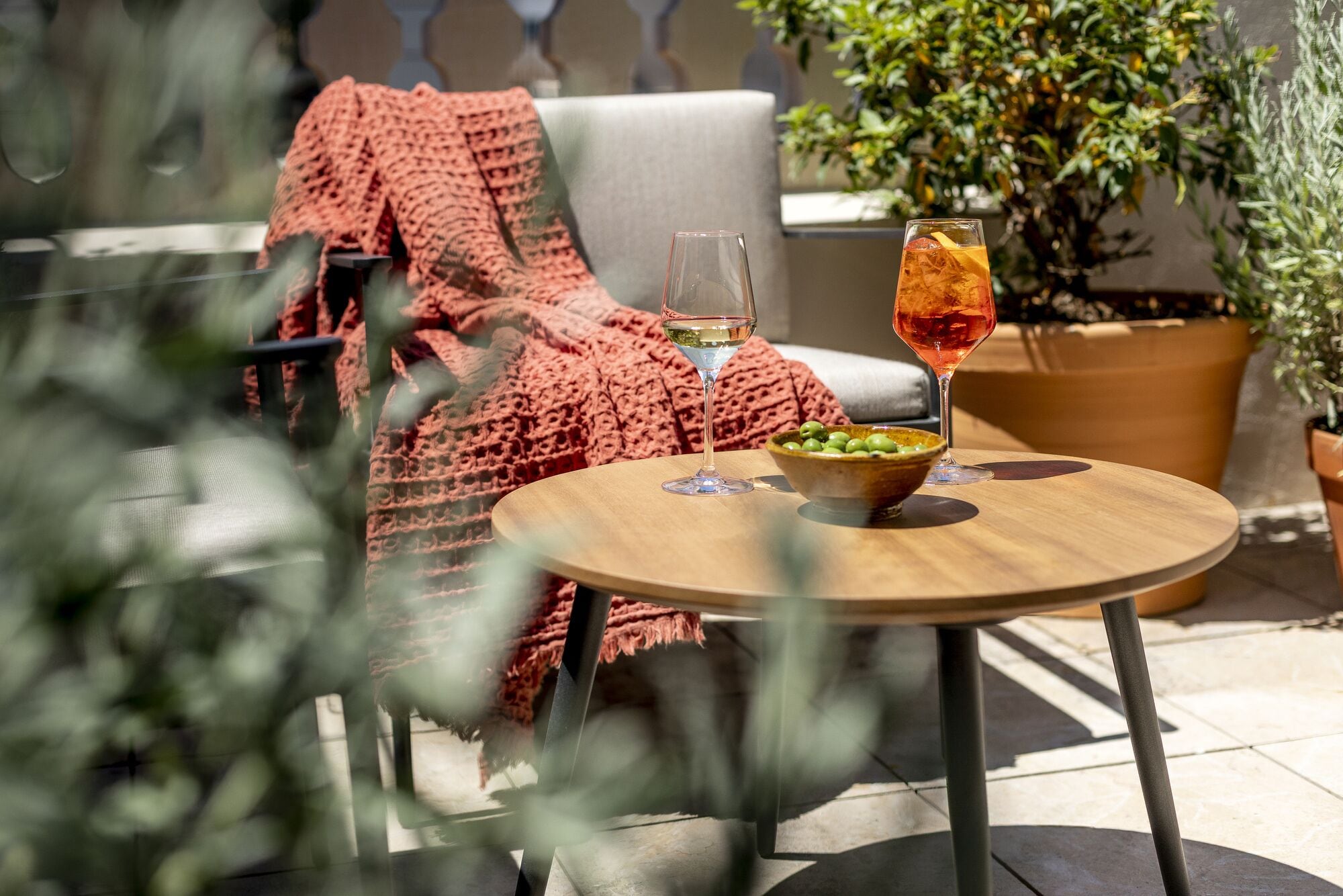 Une table ronde en bois, sur laquelle se trouvent un bol d’olives et deux verres de vin, une couverture rouge tricotée et, au fond, une plante en pot.